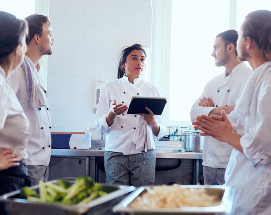 Chef holding a tablet speaking to other chefs in a kitchen.