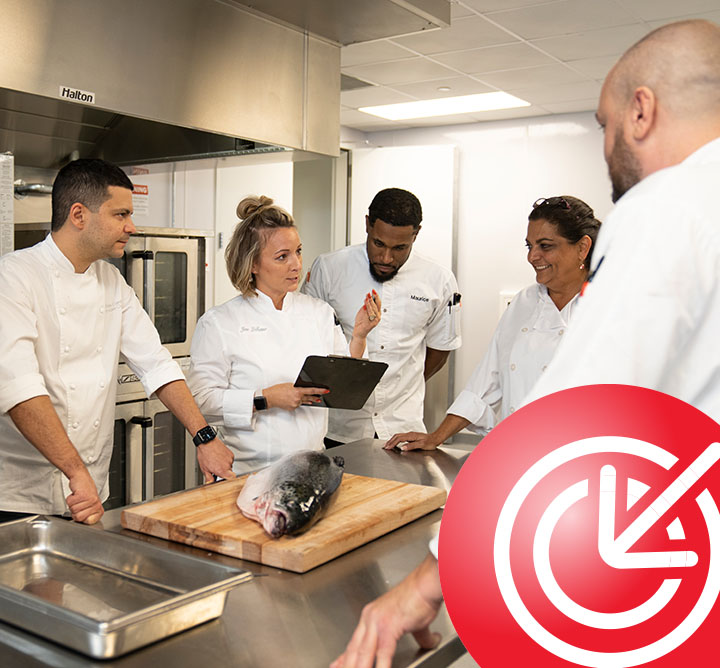 A female chef holding a tablet gives directions to other chefs in a commercial kitchen.