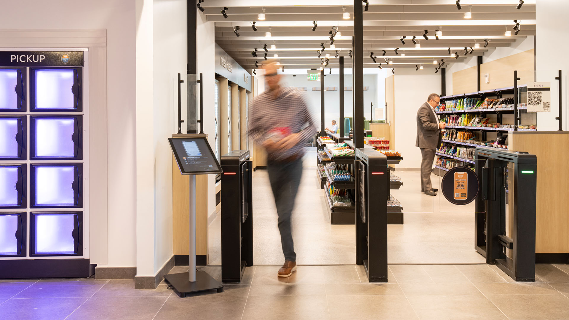 A man exiting the automated gate of a snack and beverage shop while another man browses.