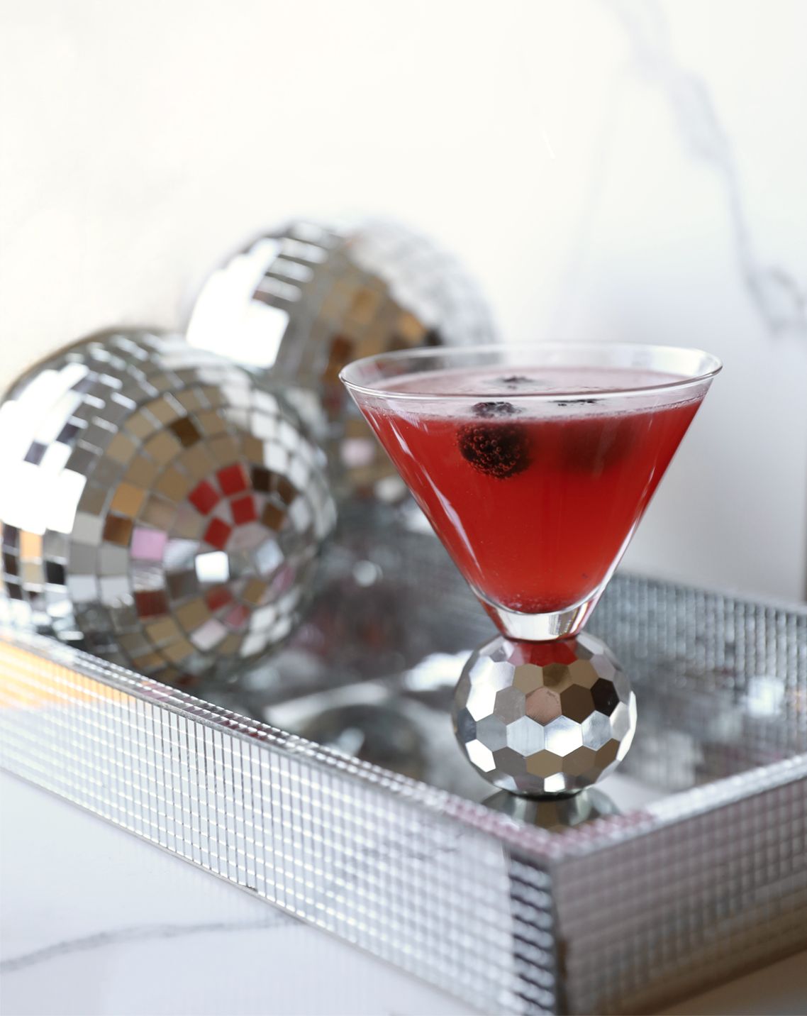 A close-up of a martini glass filled with a pink cocktail. The drink is garnished with fresh blackberries and is resting on a silver tray with disco balls.