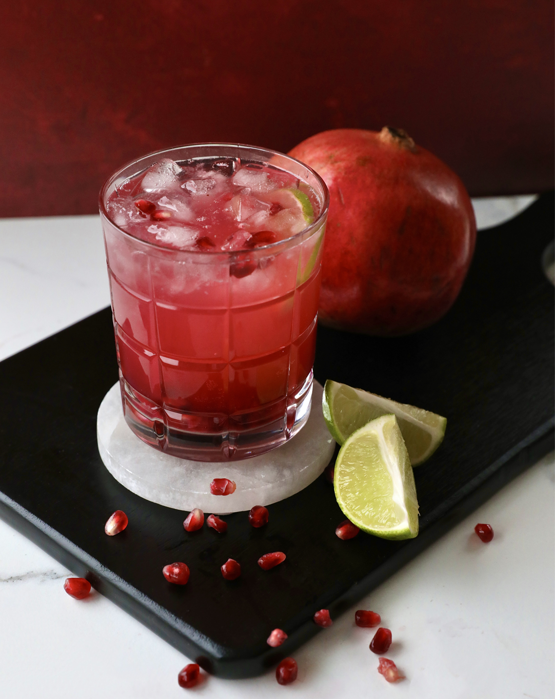 A glass of pink cocktail with ice, garnished with lime wedges and pomegranate seeds. A whole pomegranate and lime wedges are placed on a cutting board behind the glass.