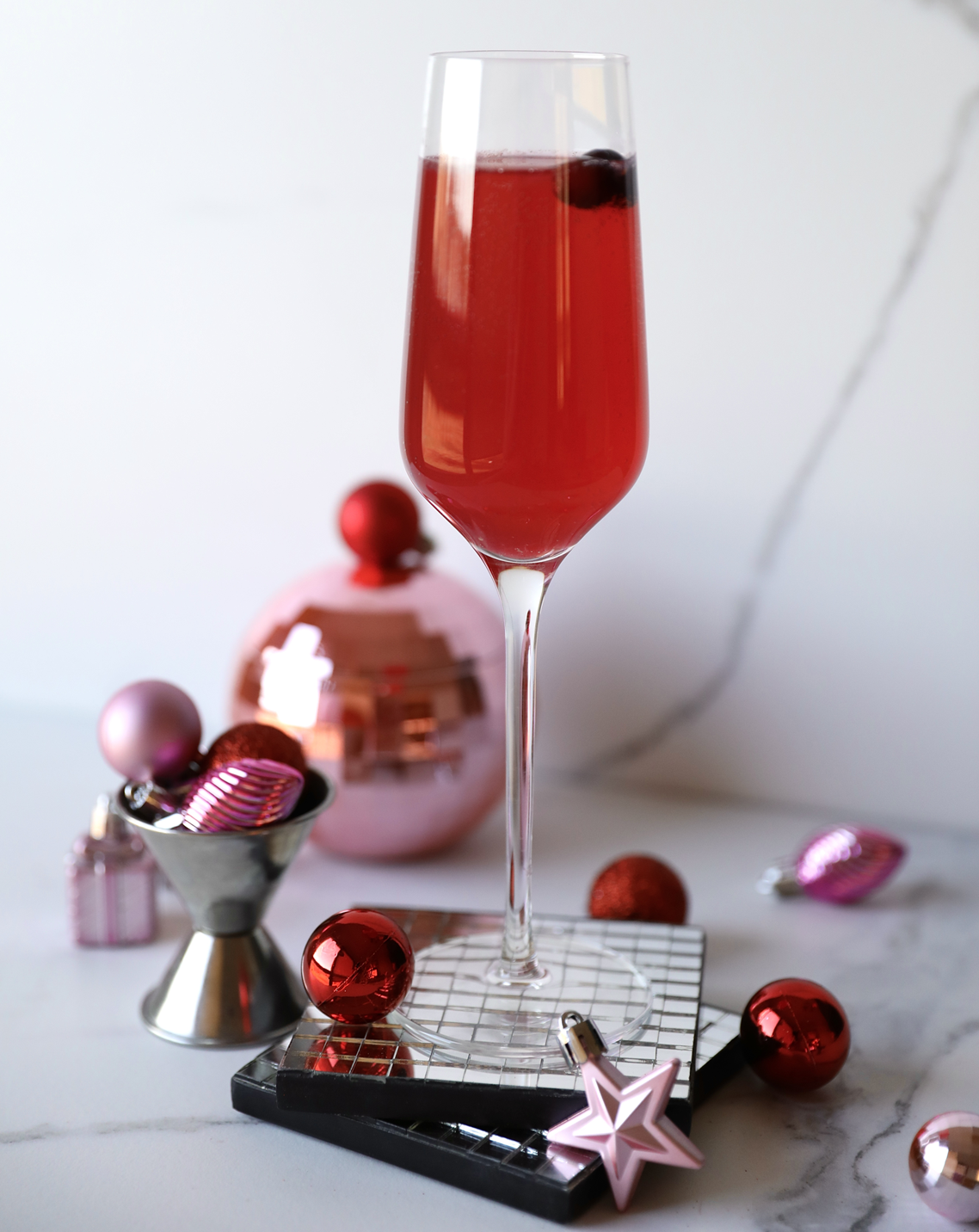A tall glass filled with a pink cocktail garnished with a cranberry. The glass is surrounded by small, colorful Christmas ornaments.