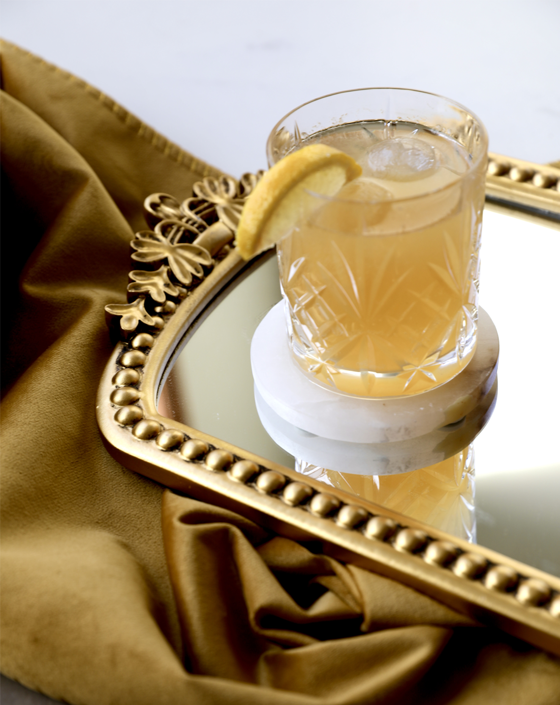 A glass of a gold drink with ice and a lemon slice on a marble coaster. The glass is on a gold tray with a gold cloth in the background.