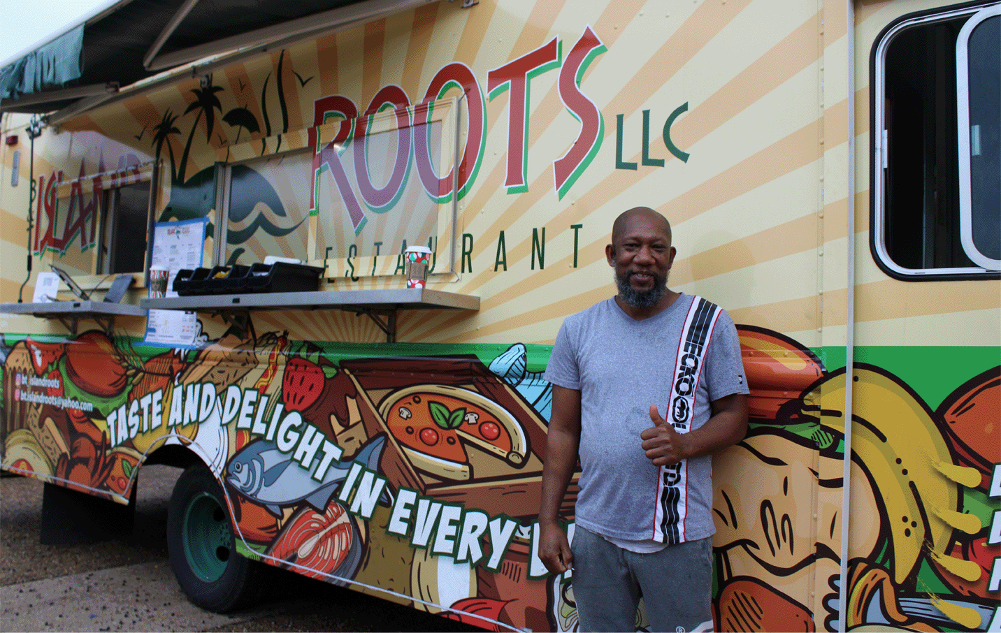 A smiling man gives a thumbs up in front of an Island Roots food truck.