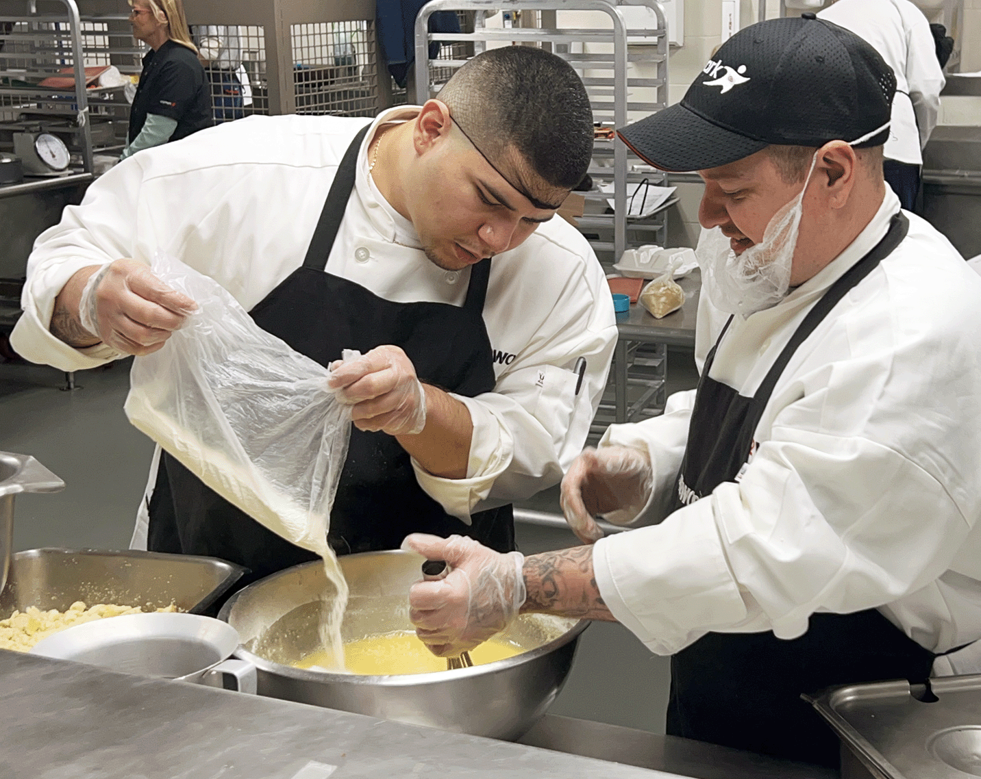 Two IN2WORK participants in a professional kitchen are mixing ingredients. One participant pours a cake mix from a bag into a bowl while the other watches.