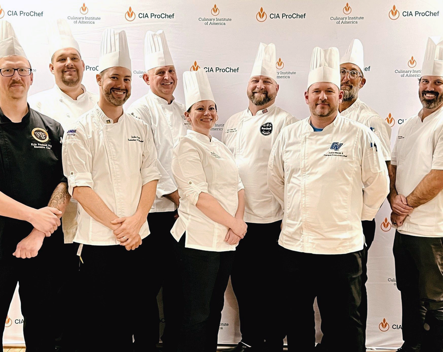 Group portrait of nine culinary professionals standing in front of a Culinary Institute of America (CIA) ProChef branded backdrop. Most are wearing white chef coats and tall white chef hats, while one person wears a black chef jacket. The group is arranged in two rows and facing the camera at an indoor event or professional gathering. The backdrop features repeated CIA ProChef and Culinary Institute of America logos.