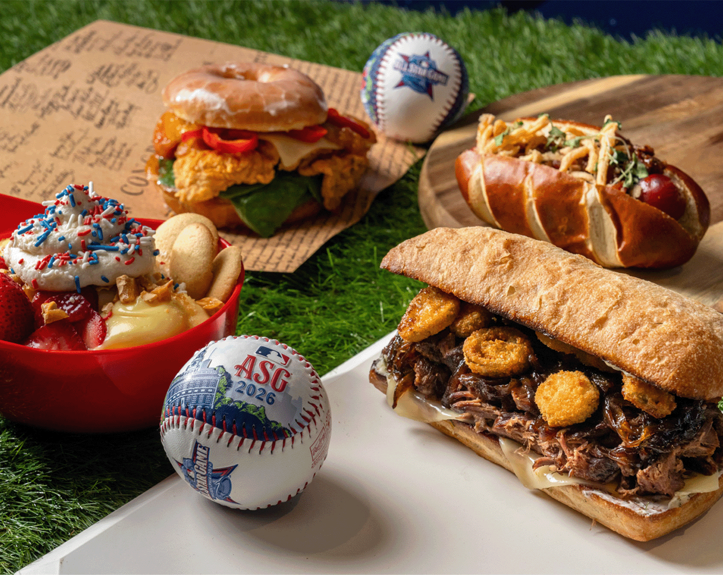 Colorful ballpark-style spread on artificial turf featuring a glazed donut fried chicken sandwich on paper, a loaded hot dog on a wooden board, a short rib sandwich topped with onion rings on a white plate, and a dessert bowl with strawberries, vanilla wafers, whipped cream, and red, white, and blue sprinkles. Two baseballs with “ASG 2026” and All-Star Game graphics sit among the food.