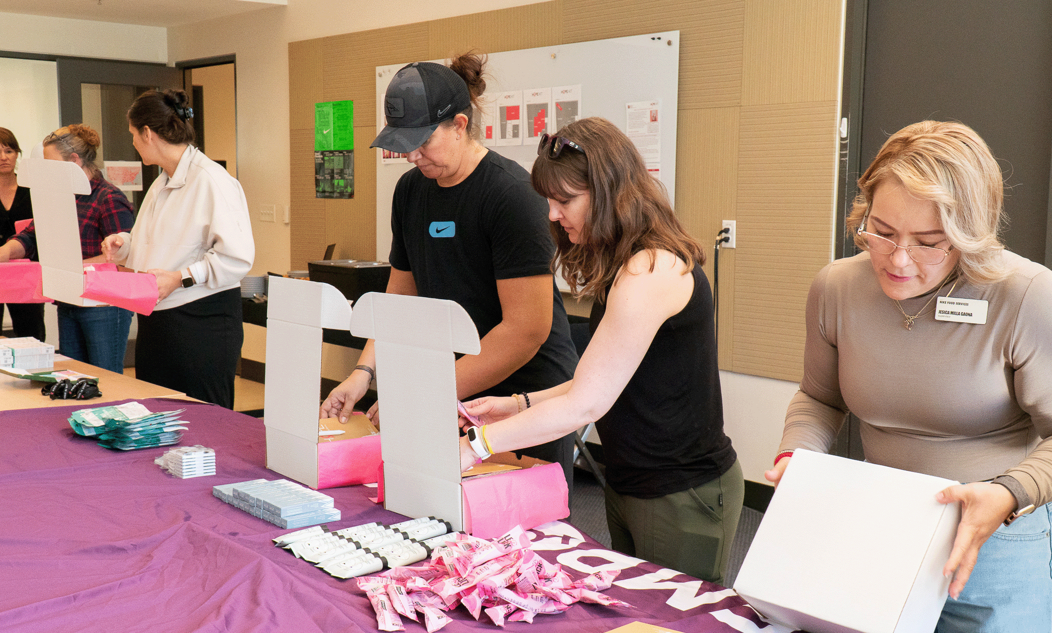 Several volunteers assemble pink-themed care packages at a table covered with supplies, folding white boxes and packing personal care items during a community service event in a meeting room.