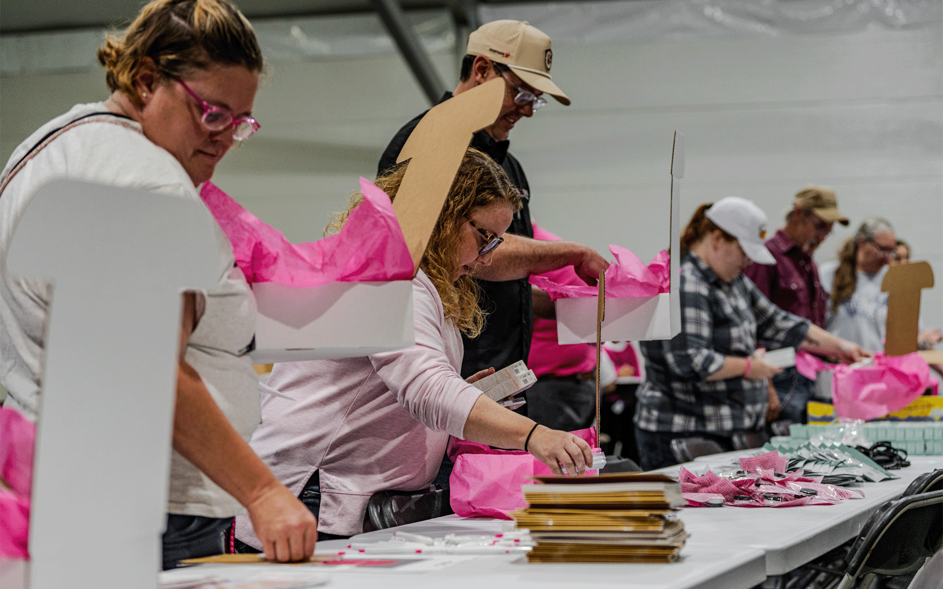Volunteers stand side by side at a long table assembling HOPE Kits, placing personal care items and resource materials into boxes lined with bright pink tissue paper. Supplies, informational booklets, and completed kits are arranged across the table during a community service event.
