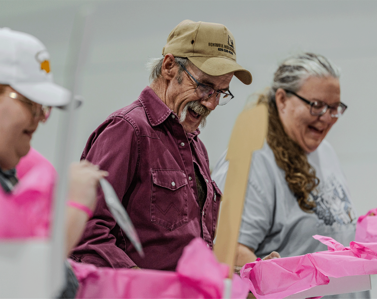 Volunteers assemble HOPE Kits at a community service event, working around tables filled with pink tissue-lined boxes and care package supplies. Several participants sort materials and pack items into boxes as part of a breast cancer support initiative.