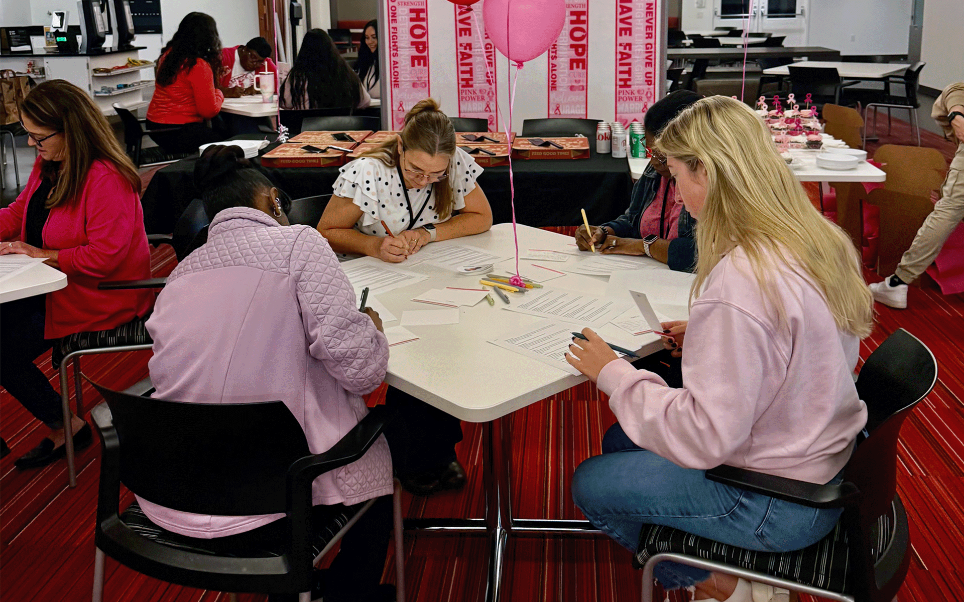 Employees sit around tables writing notes and assembling encouragement cards during a breast cancer awareness volunteer event. Pink balloons, awareness-themed banners, and refreshments are displayed throughout the room as participants work together on the activity.