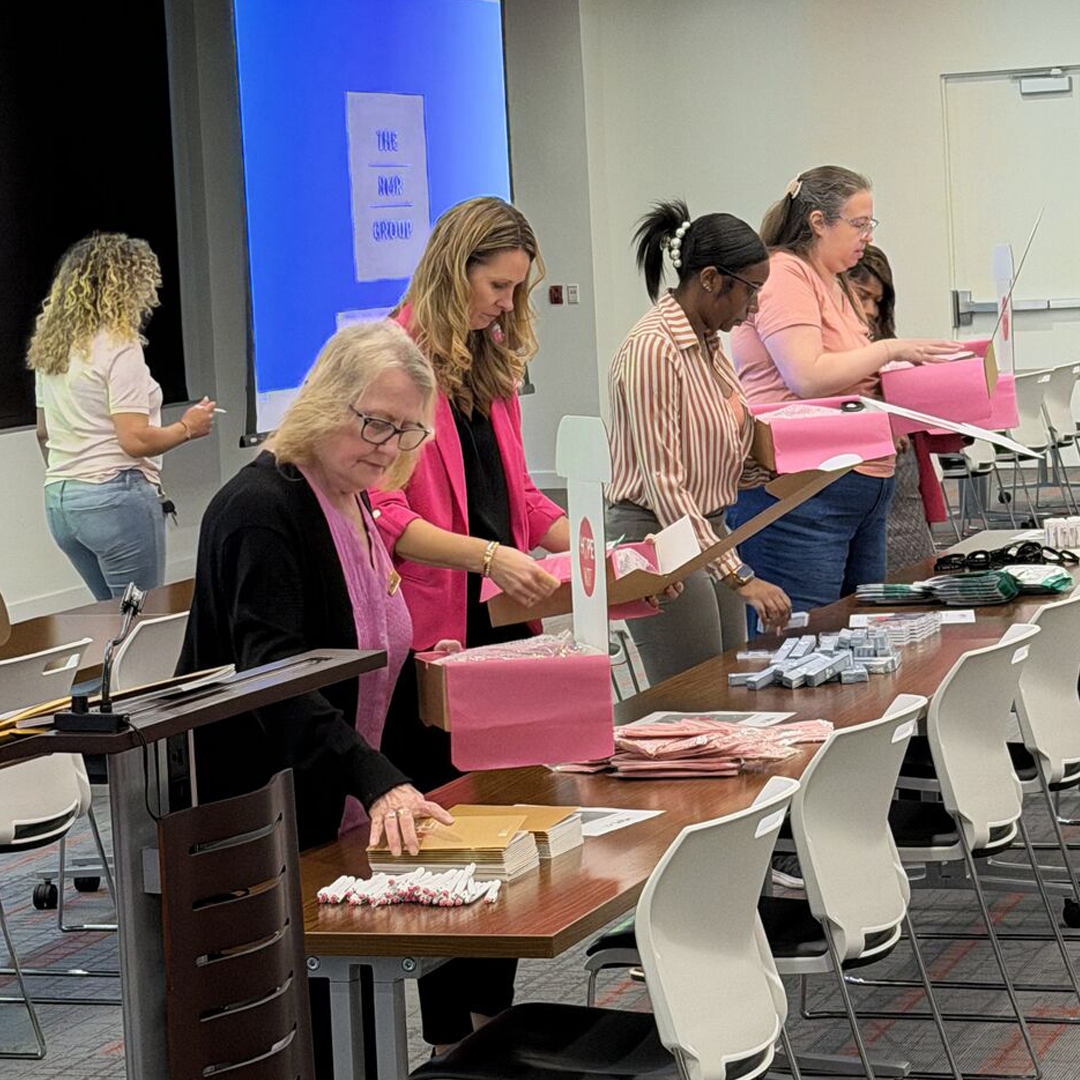 Volunteers assemble care packages at a long table in a conference room, placing personal care items, pens, and informational materials into pink tissue-lined boxes. Stacks of supplies are arranged across the table as participants work together during a community service project.