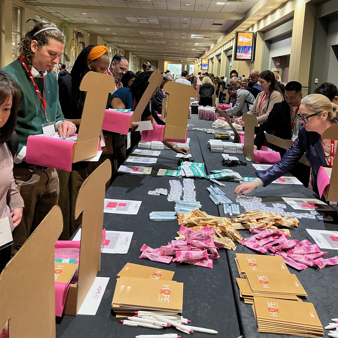 Volunteers stand on both sides of a long table assembling care packages during a large-scale community service event. Open cardboard boxes lined with pink tissue paper, personal care items, pens, and booklets labeled “HOPE” are arranged across the table as participants work together in a convention center hallway.