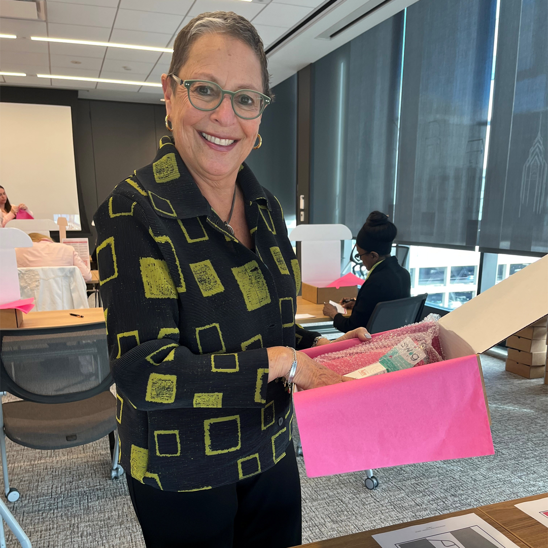 A volunteer holds an open HOPE Kit box lined with pink tissue paper and filled with personal care items during a community service event. Additional volunteers and partially assembled kits are visible in the conference room background.
