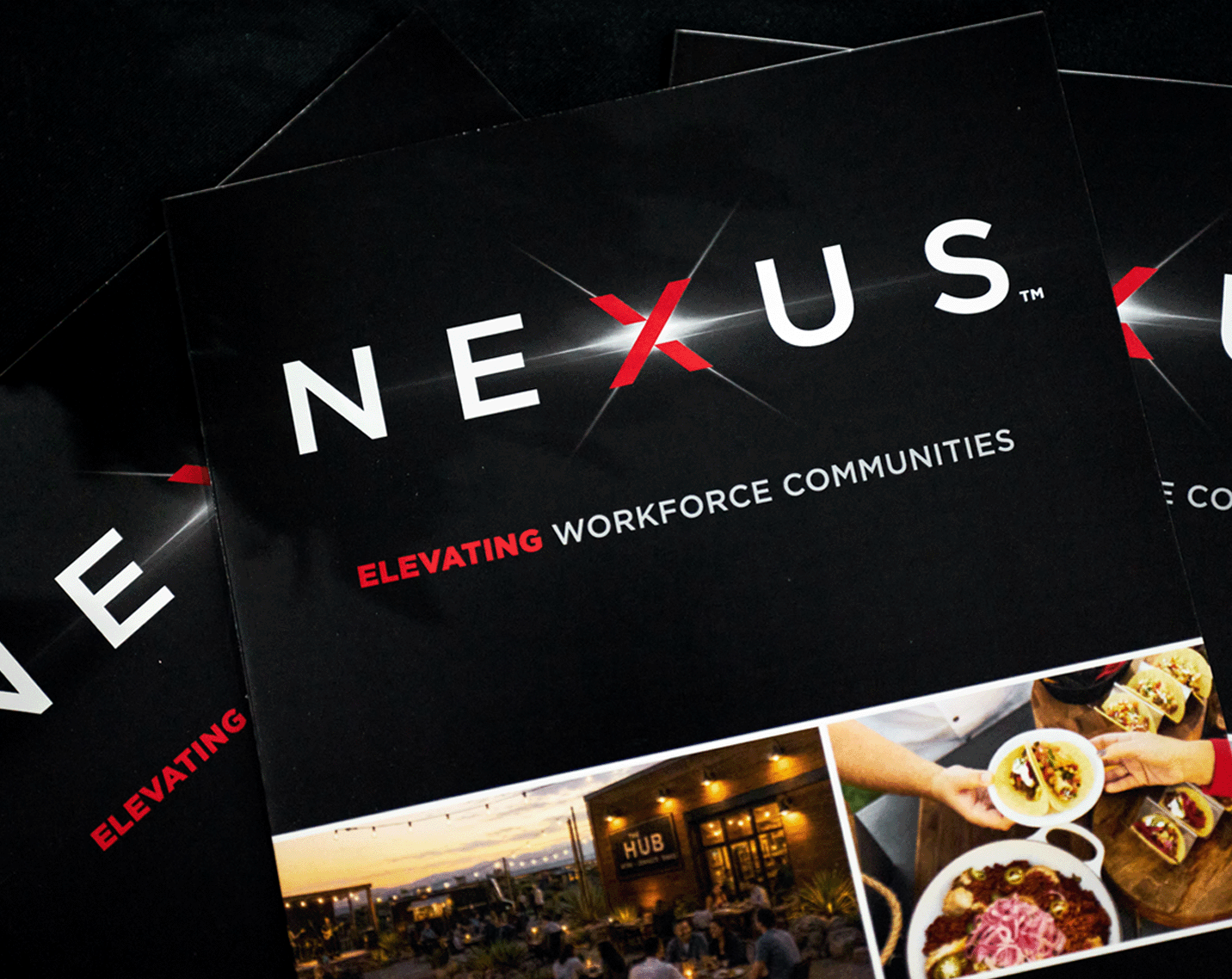 Cover of a NEXUS promotional brochure on a black background. The word “NEXUS” appears in large white letters with a red “X,” above the tagline “ELEVATING WORKFORCE COMMUNITIES.” Along the bottom are photos showing an outdoor gathering space, a building labeled “HUB,” and plated food being served. Additional copies of the brochure are partially visible behind the main brochure.