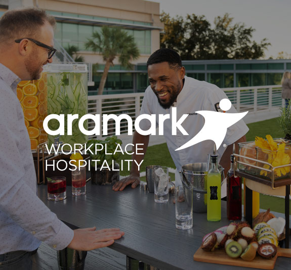 Guest and server standing at a table with fruit-infused drinks and a fresh food display, with the Workplace Hospitality logo.
