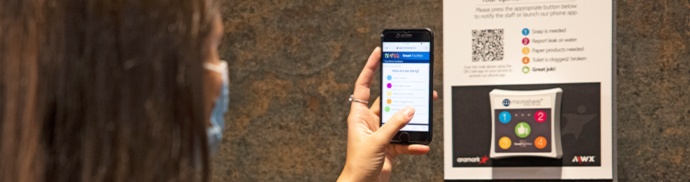 A masked corporate employee using her phone to scan a QR code on a digital feedback device.