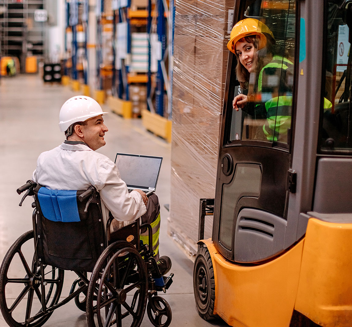 A man in a wheelchair wearing a white hardhat talking to a women driving a forklift and wearing a hardhat and safety gear.