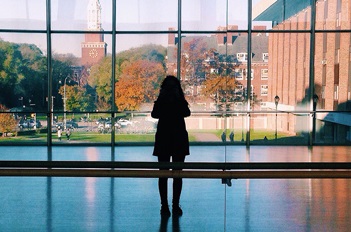 A person looking out large bay windows at a campus landscape.