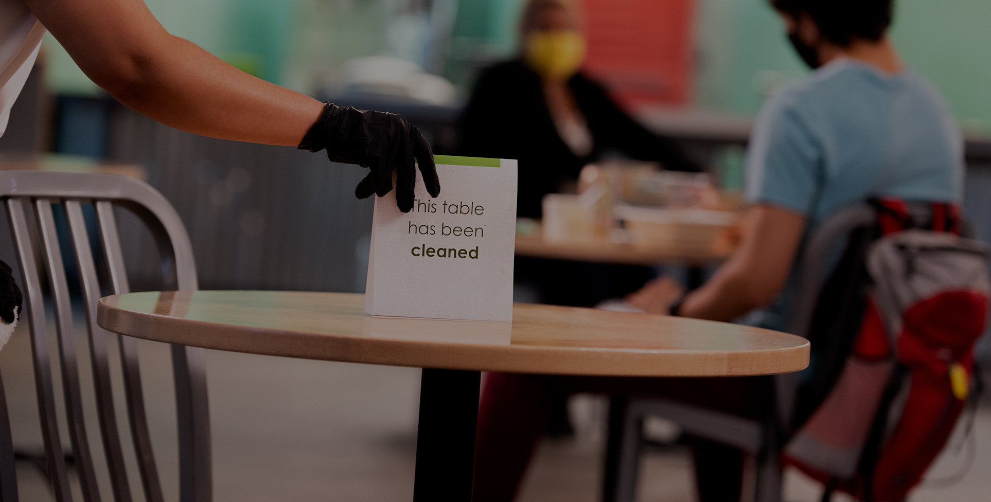 In a campus dining hall, a worker puts a sign on a table stating it's been cleaned.