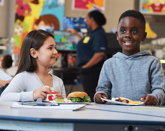 A young girl and boy with food in a school cafeteria.
