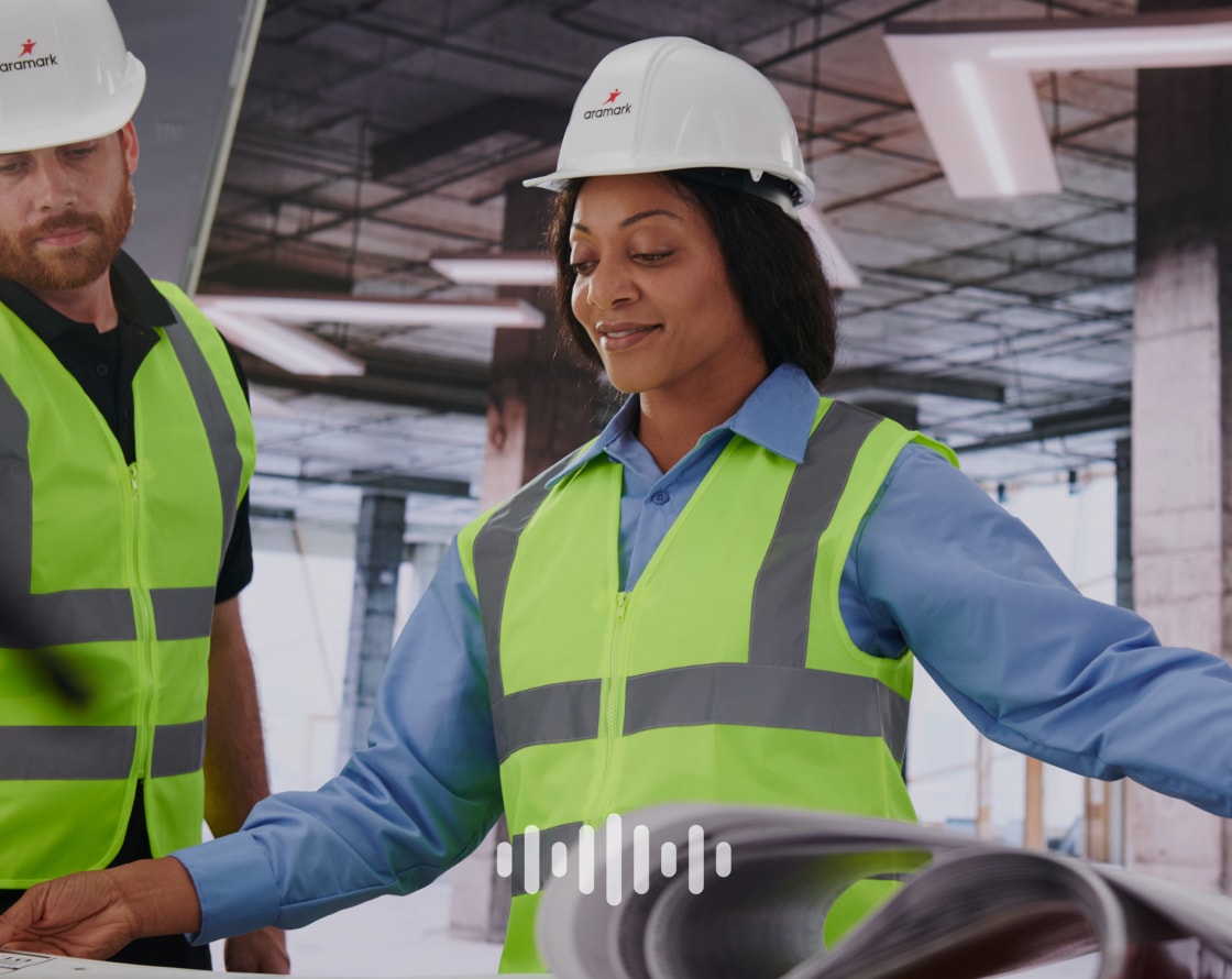 Workers wearing Aramark hard hats and high visibility vests looking at construction plans, and an overlaid audio symbol.
