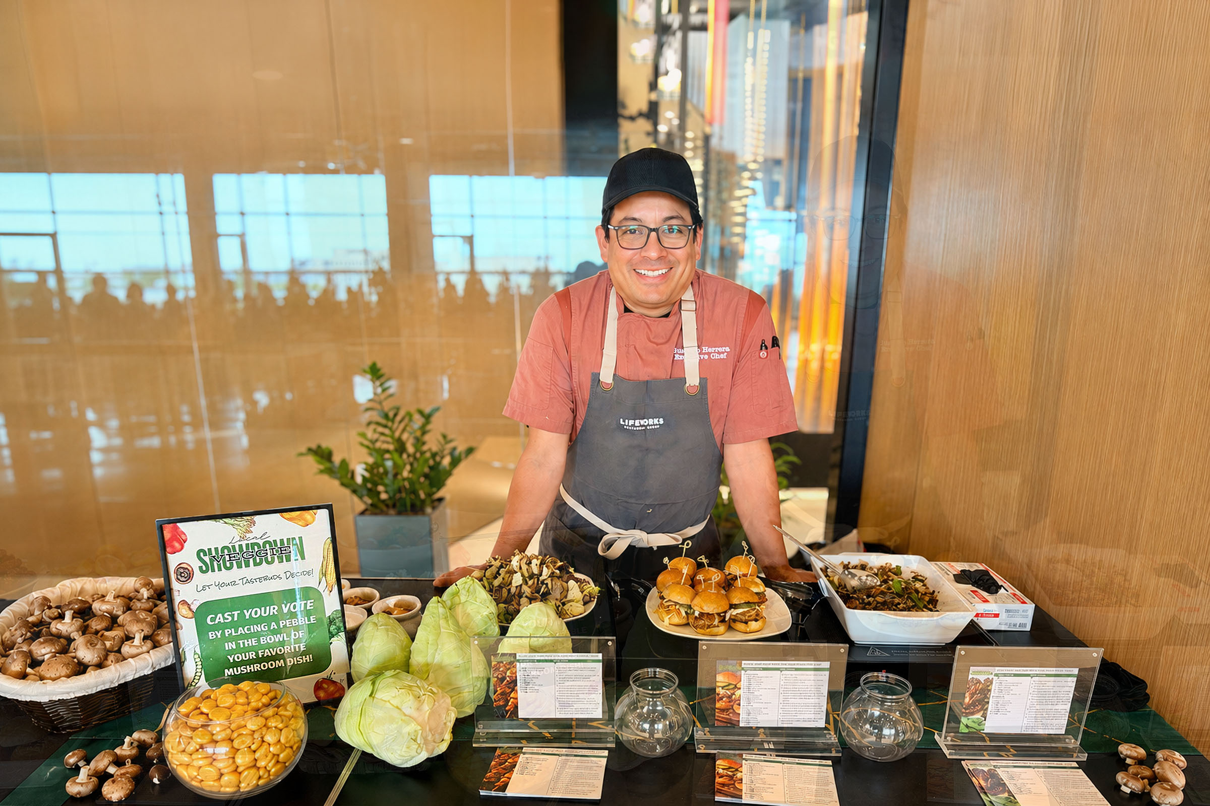 LifeWorks Chef smiling over a table of food samples and a sign to cast a vote for your favorite dish.