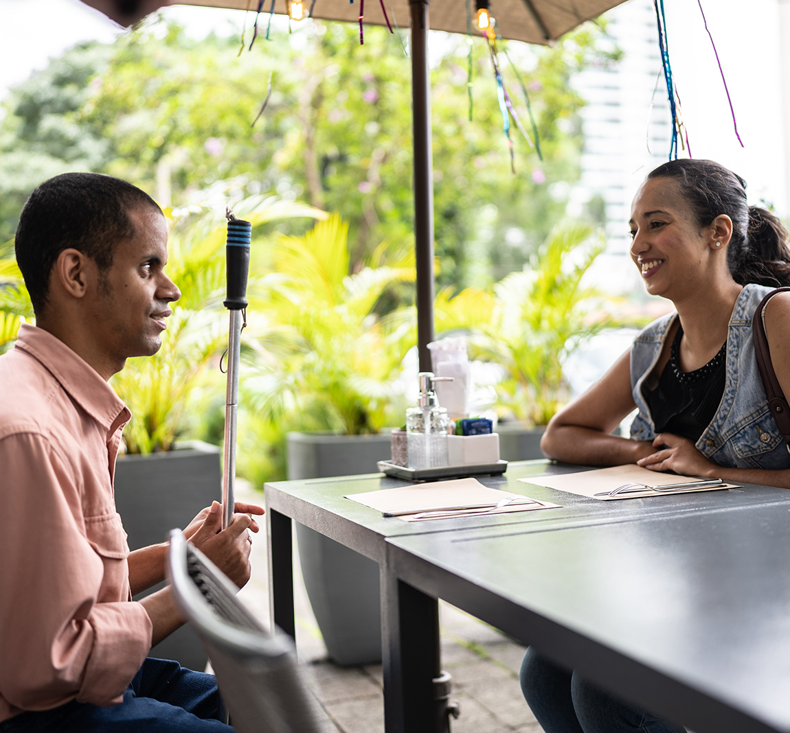 A woman and a man holding a cane smile at each other across the table of an outdoor restaurant.