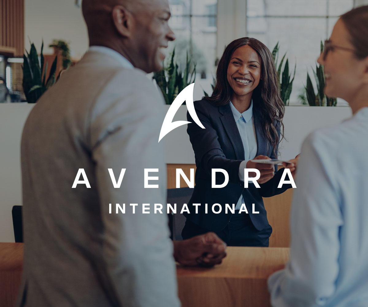Woman at desk handing a hotel key to guest with Avendra logo. 