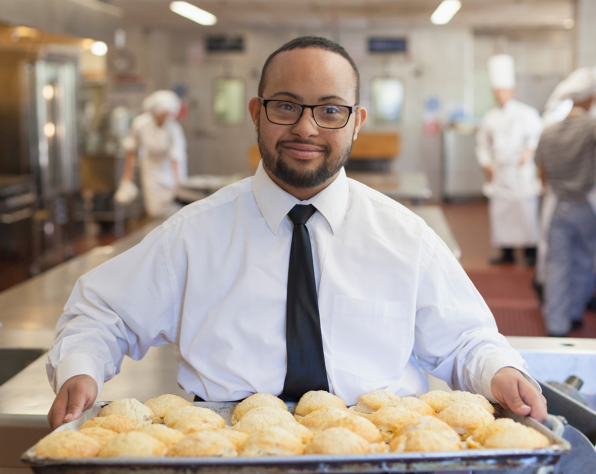 A young man in a button-down shirt and tie carrying a tray of freshly baked rolls.
