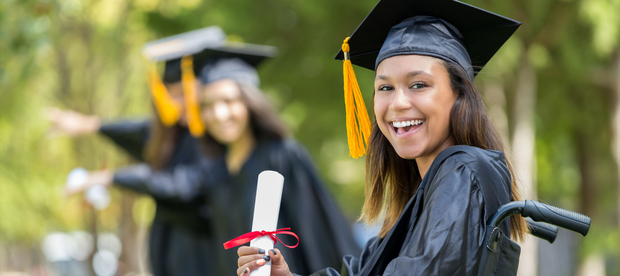 A young woman dressed in a black tasseled cap and gown sits in a wheelchair holding up a wrapped diploma.