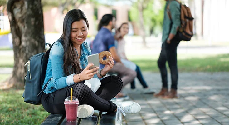 Student sitting on a bench with a donut and smoothie, looking at her phone.