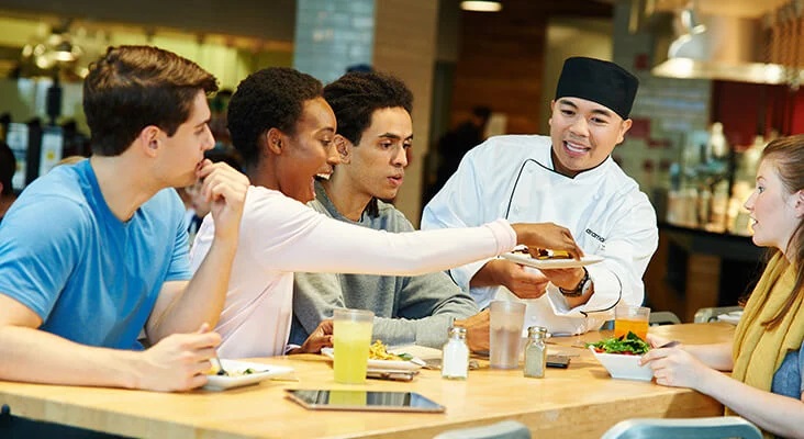 An Aramark chef offers food on a plate to students sitting together in a dining hall.