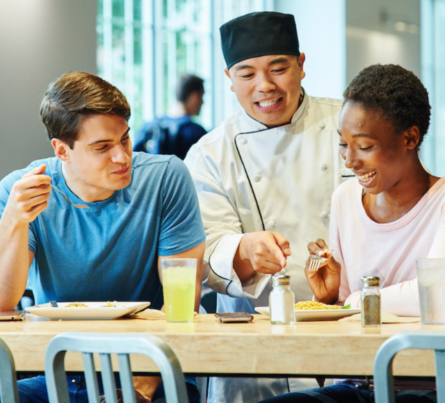 Chef talks to two students about their food while they eat.