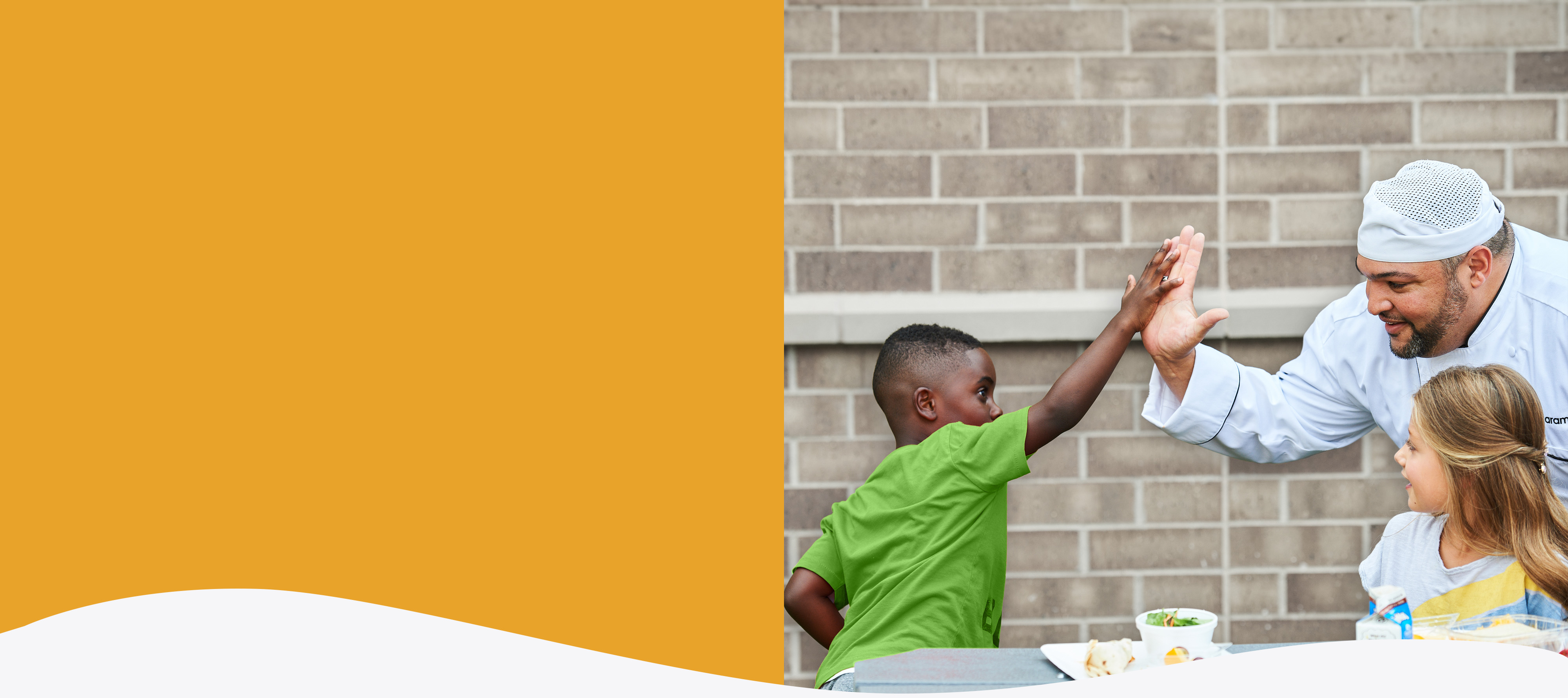 A young child sitting with a tray of food giving a high five to a smiling Aramark chef.