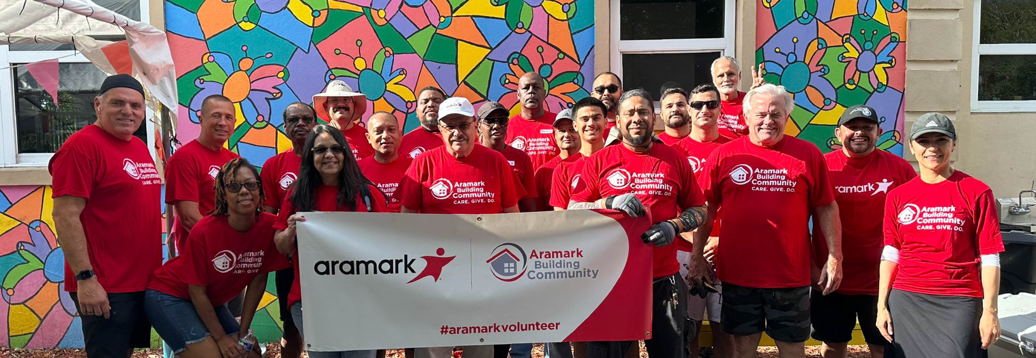 A group of smiling volunteers wearing Aramark Building Community shirts and holding up a banner.