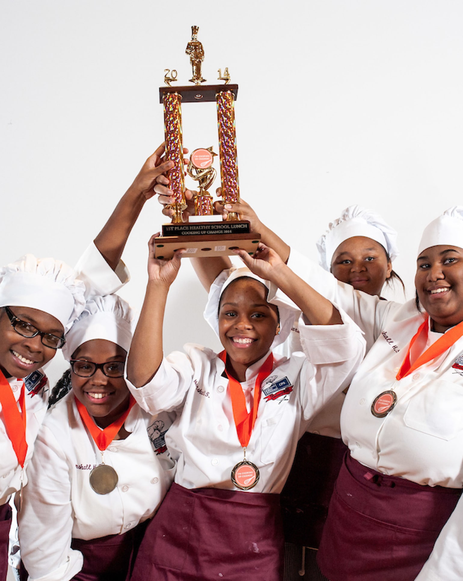 Young women in white chef's jackets and hats triumphantly raising up a trophy and showing off the medals around their necks.