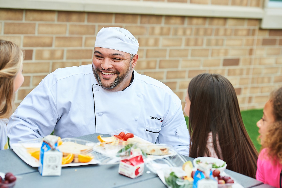 Chef sitting at a lunch table talking to elementary school students.