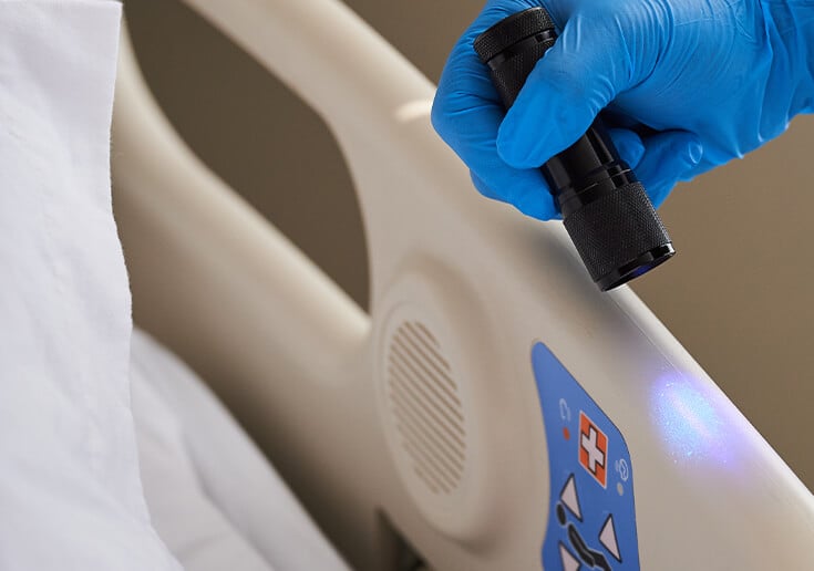 A worker using a fluorescent gel and a black light to inspect the sanitization of a hospital bed.