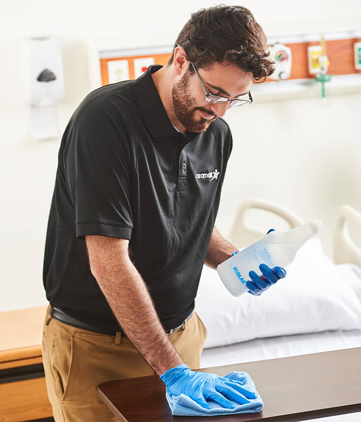 An Aramark employee wearing latex gloves wiping down surfaces of a hospital room with disinfectant solution.