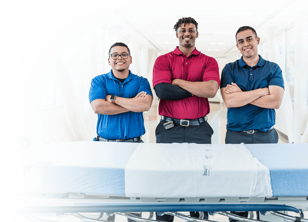 Smiling healthcare workers with their arms crossed standing behind an empty hospital bed.