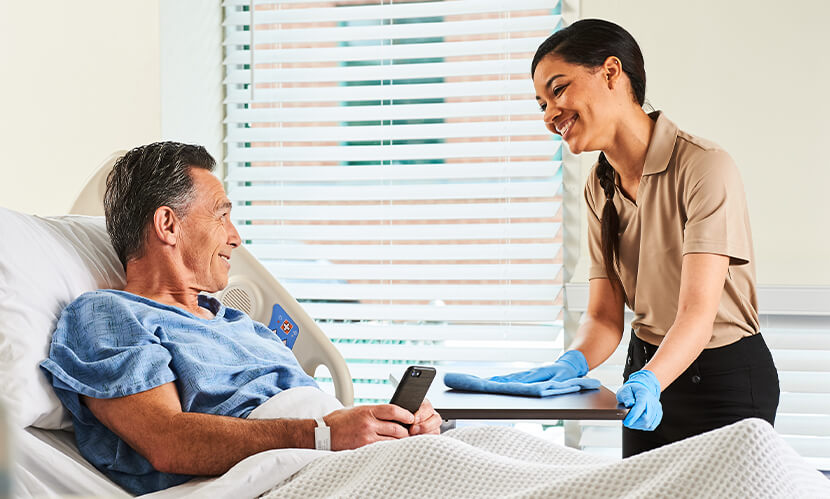 A hospital employee wipes down a bedside tray table while talking with a patient.