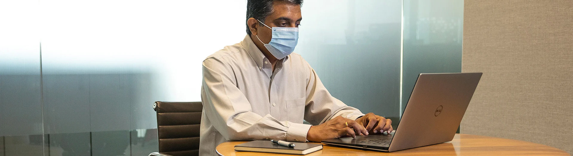 A masked corporate employee sitting at a desk, working on a laptop.