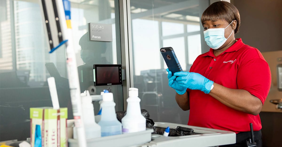 An Aramark worker wearing PPE stands outside of an office with a cleaning cart, using her cell phone.
