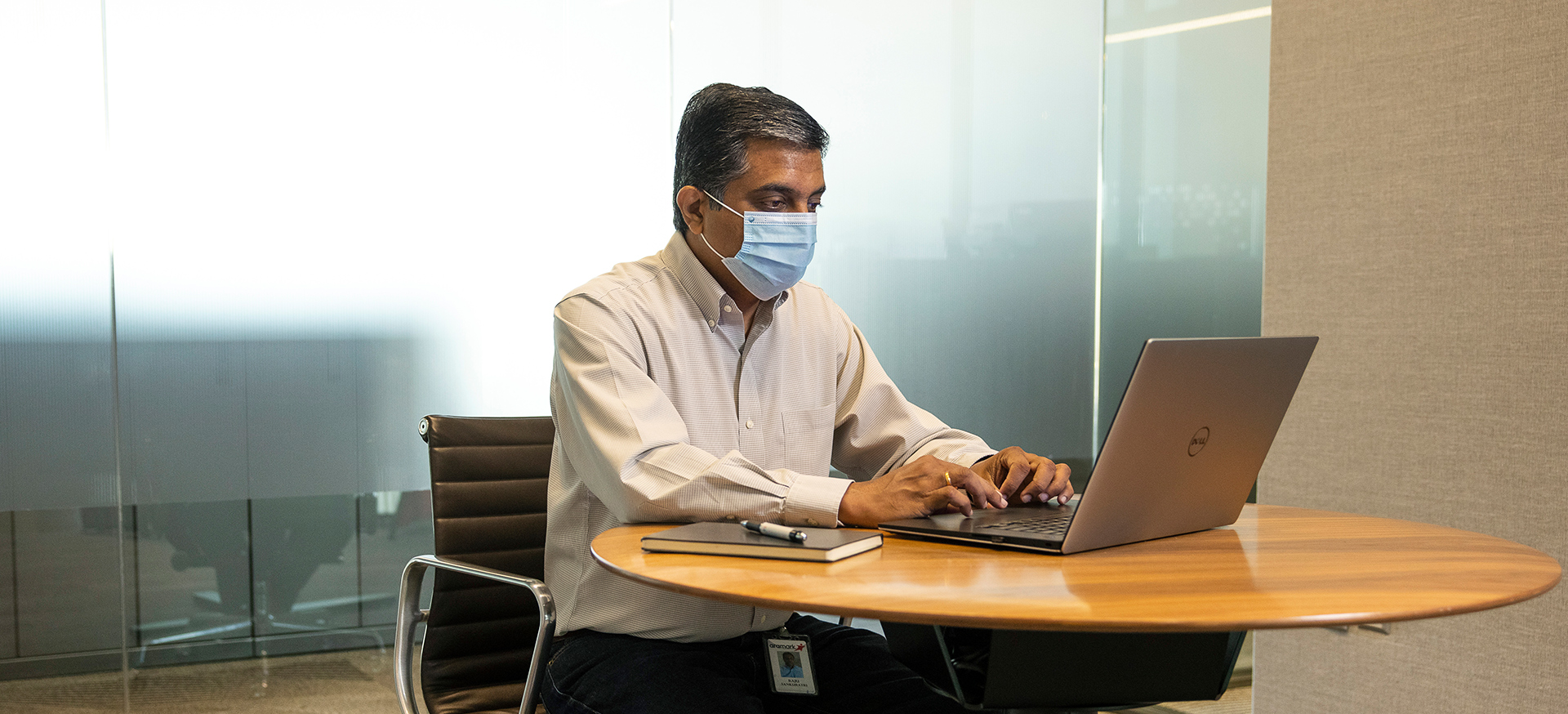 A masked corporate employee sitting at a desk, working on a laptop.
