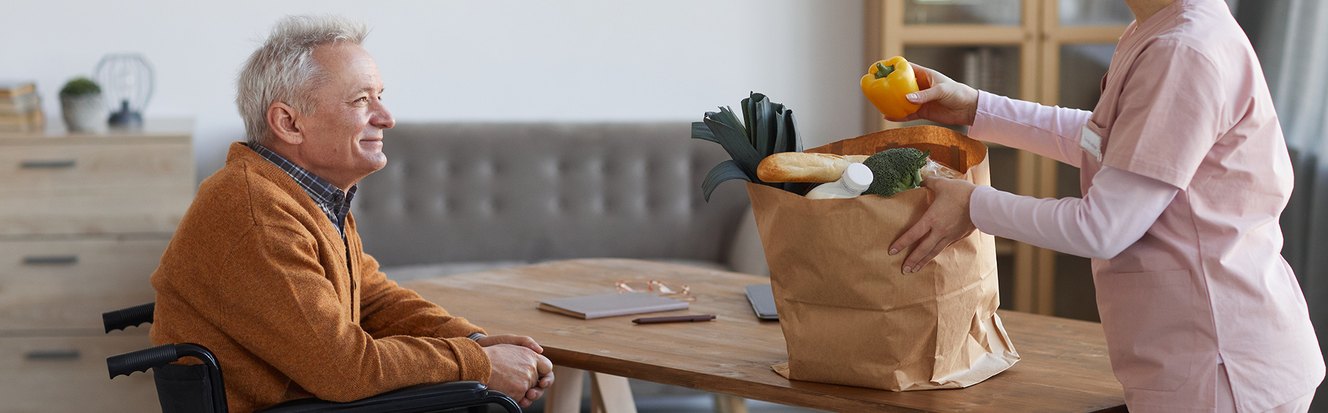 Older man in a wheelchair with an aide carrying a bag of groceries.
