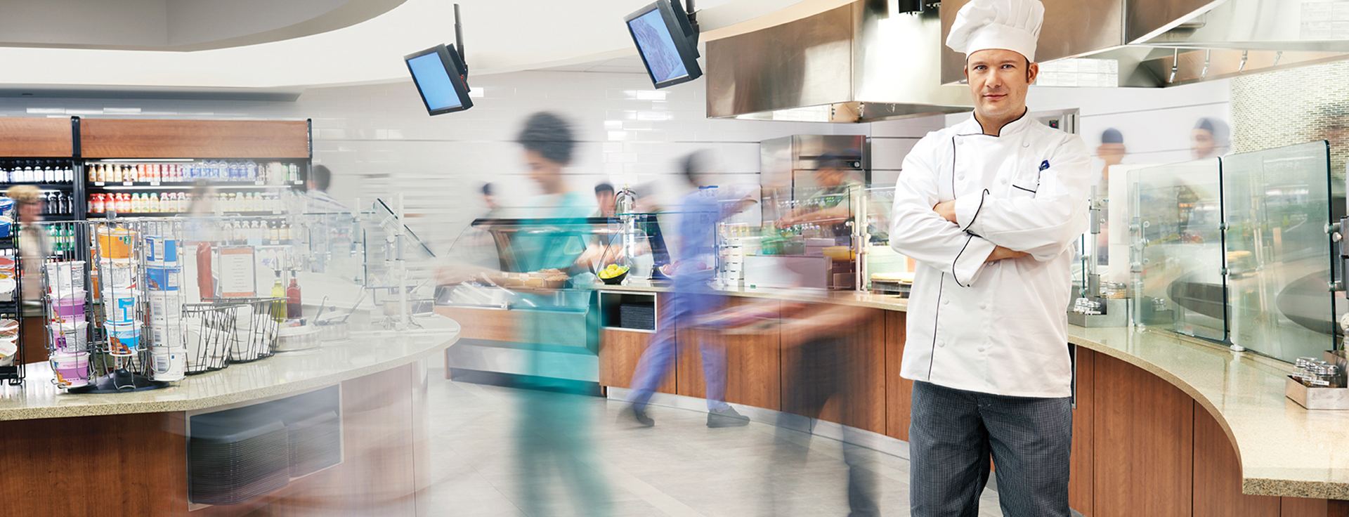Chef standing in a hospital cafeteria while nurses and doctors pass by.