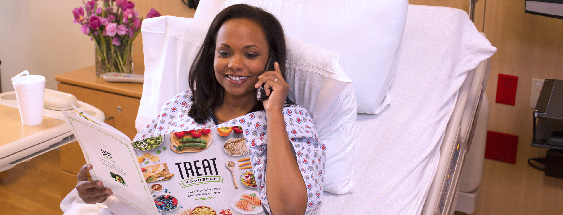 Female patient in a hospital bed using a phone and looking at a hospital food delivery menu.