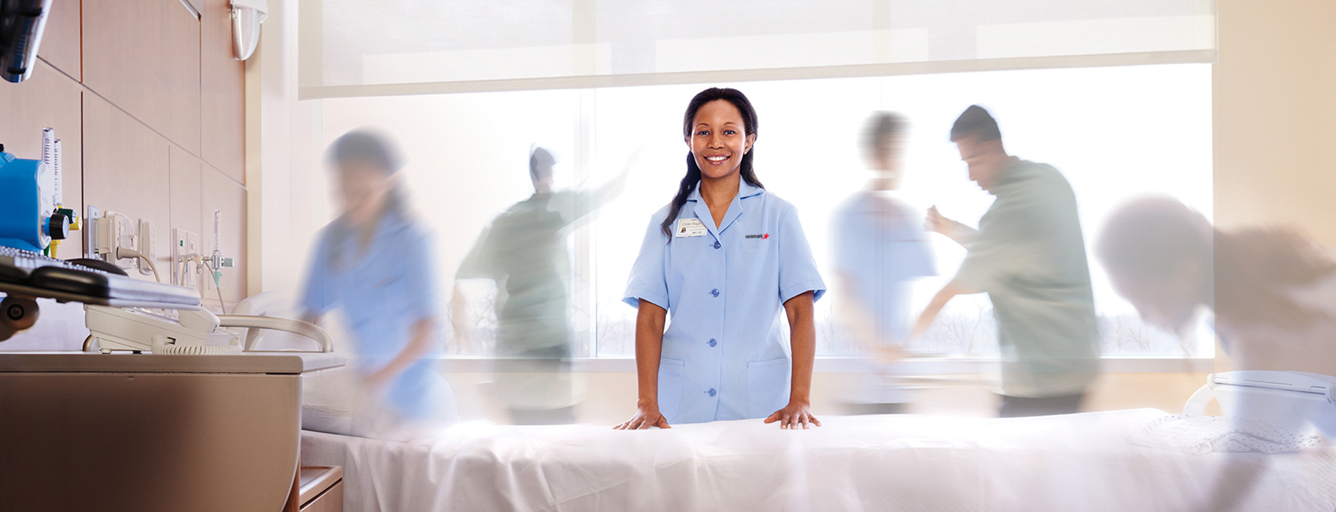 An Aramark worker stands smiling at a hospital bed while EVS employees clean the room.