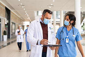 Doctor and nurse wearing masks walking and discussing the information on a clipboard.